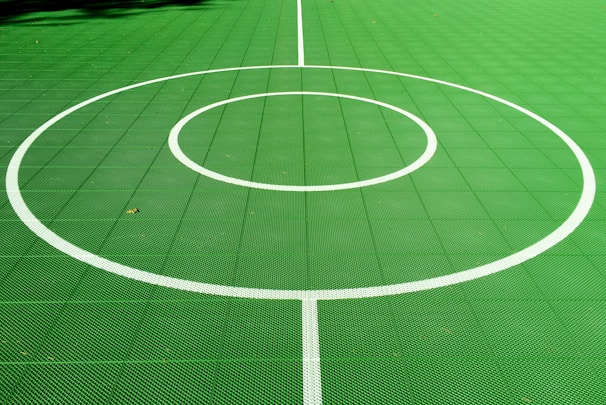 A vibrant green sports court featuring distinct white circular markings and straight lines. The court surface is composed of a textured grid pattern, and small leaves can be seen scattered on the surface.