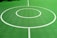 A vibrant green sports court featuring distinct white circular markings and straight lines. The court surface is composed of a textured grid pattern, and small leaves can be seen scattered on the surface.
