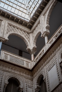 Intricate architectural details of an arched building interior feature ornate carvings and designs, emphasizing classical and historical aesthetics. Arched windows and columns support a decorative ceiling with a glass roof that filters light into the space.