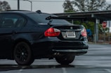 A black BMW 328i with a visible rear wing parked in a wet parking lot. The car has visible raindrops and a California license plate. In the background, a stop sign and some leafless trees can be seen.