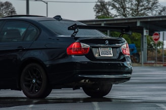 A black BMW 328i with a visible rear wing parked in a wet parking lot. The car has visible raindrops and a California license plate. In the background, a stop sign and some leafless trees can be seen.