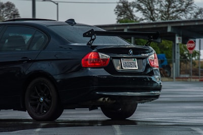 A black BMW 328i with a visible rear wing parked in a wet parking lot. The car has visible raindrops and a California license plate. In the background, a stop sign and some leafless trees can be seen.