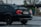 A black BMW 328i with a visible rear wing parked in a wet parking lot. The car has visible raindrops and a California license plate. In the background, a stop sign and some leafless trees can be seen.