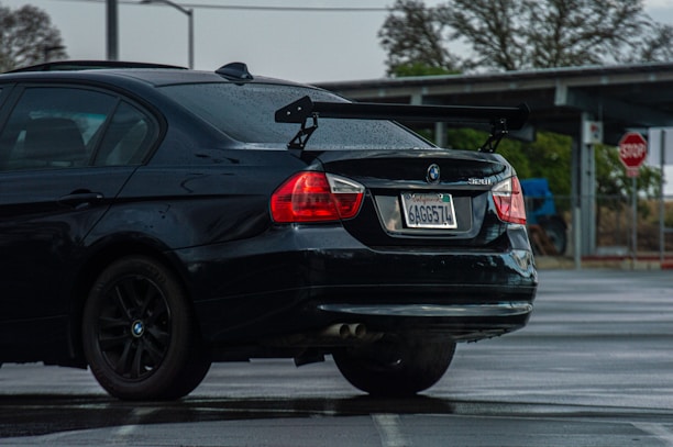A black BMW 328i with a visible rear wing parked in a wet parking lot. The car has visible raindrops and a California license plate. In the background, a stop sign and some leafless trees can be seen.