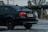 A black BMW 328i with a visible rear wing parked in a wet parking lot. The car has visible raindrops and a California license plate. In the background, a stop sign and some leafless trees can be seen.