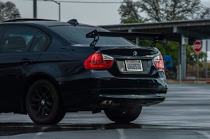 A black BMW 328i with a visible rear wing parked in a wet parking lot. The car has visible raindrops and a California license plate. In the background, a stop sign and some leafless trees can be seen.