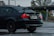 A black BMW 328i with a visible rear wing parked in a wet parking lot. The car has visible raindrops and a California license plate. In the background, a stop sign and some leafless trees can be seen.