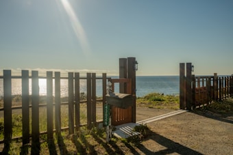 A scenic view of the ocean is framed by a wooden fence and gate. Sunlight glistens on the water's surface, creating a serene and inviting atmosphere. A small mailbox with a house number is visible in the foreground.