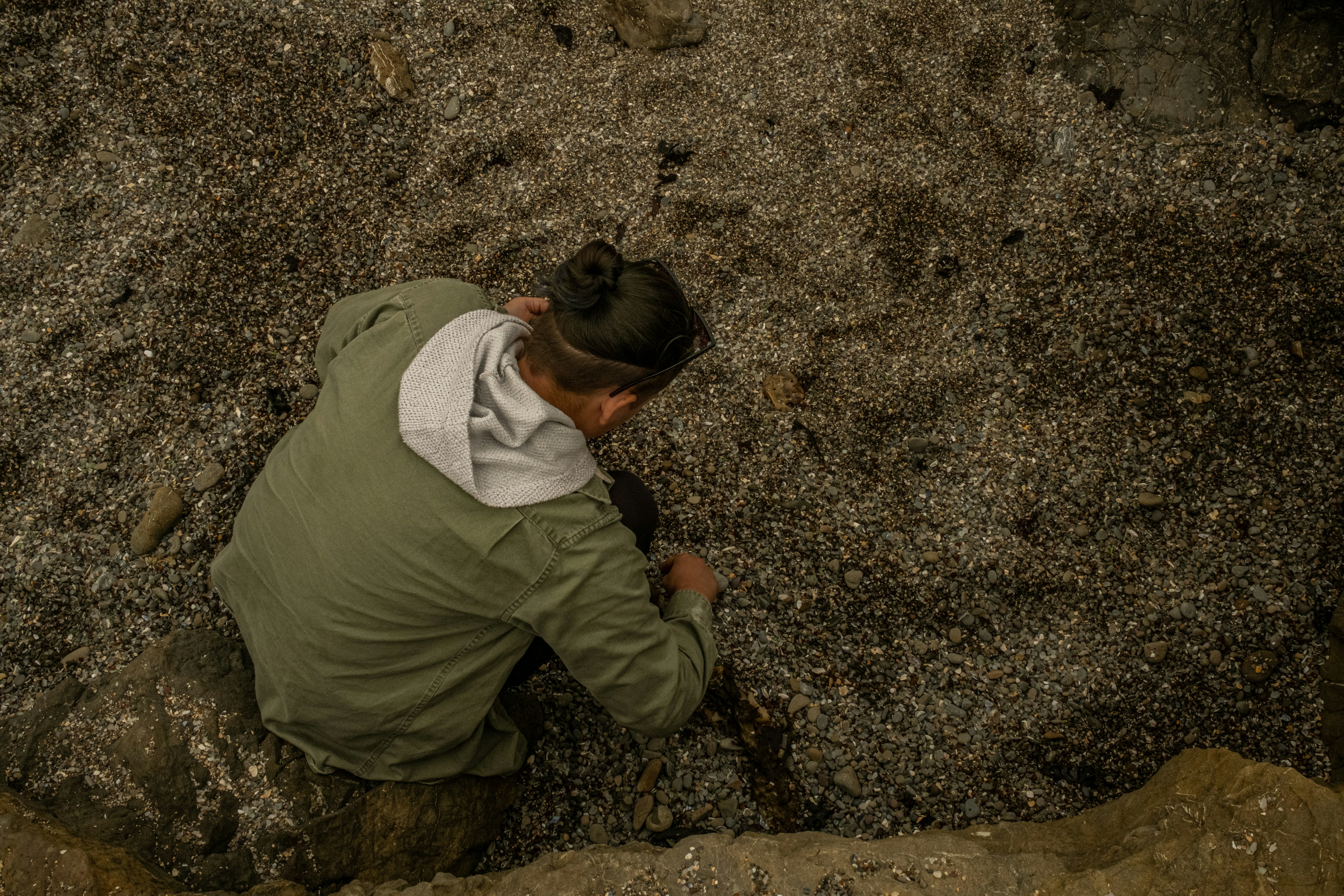 A lone figure crouches on a pebbled shoreline, wearing a green jacket with a gray hood, examining something near the rocks.