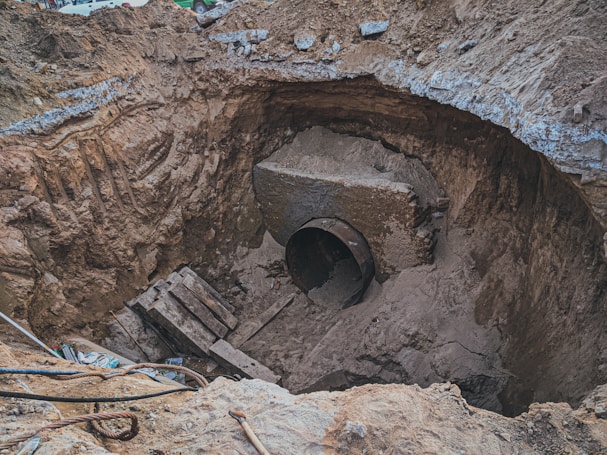 A large excavation site with a significant amount of earth removed, revealing a large, corroded metal pipe partially buried under soil. The surroundings are rough and uneven, with scattered construction debris such as wooden planks and cables. The walls of the excavation show distinct layers of soil and dirt.