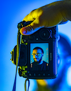 A hand holding a Canon camera with a live view screen displaying a portrait of a man wearing glasses. The background is a vibrant mix of blue and yellow tones, creating a striking contrast with the camera's black exterior.