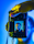 A hand holding a Canon camera with a live view screen displaying a portrait of a man wearing glasses. The background is a vibrant mix of blue and yellow tones, creating a striking contrast with the camera's black exterior.