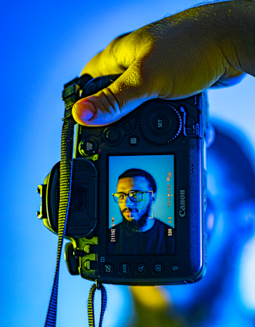A hand holding a Canon camera with a live view screen displaying a portrait of a man wearing glasses. The background is a vibrant mix of blue and yellow tones, creating a striking contrast with the camera's black exterior.
