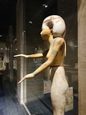 A wooden statue of ancient Egyptian style is displayed in a glass case. The statue features a figure with elongated limbs and a traditional headdress, standing with arms extended forward. The background includes stone walls and additional exhibits under gentle lighting.