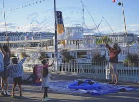 A man creates large soap bubbles using a long wand while children eagerly watch and reach for them. In the background, a large white boat is docked, with buildings and trees visible along the waterfront. The scene is lively, set during daylight with a clear sky and festive ambiance.