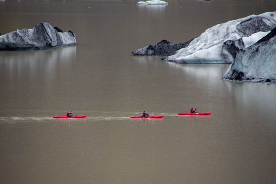 Three individuals paddle in red kayaks across a calm body of water surrounded by large gray and black rocky formations. The scene is serene, emphasizing the contrast between the vibrant kayaks and the muted tones of the water and rocks.