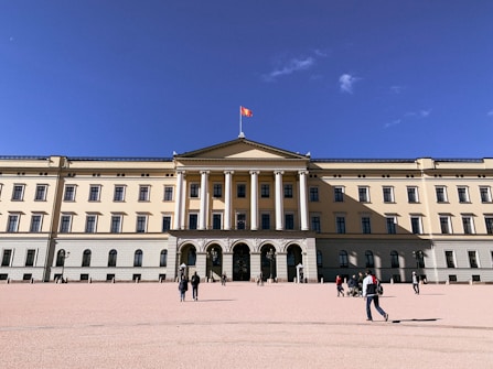 A large, neoclassical palace with several tall windows and a central pediment supported by columns. The building features a light beige facade and is topped with a flag. The foreground shows a spacious plaza with people walking and gathering around.