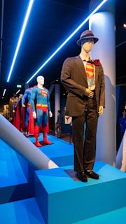Several mannequins display superhero costumes in a museum setting. The foreground features a mannequin dressed in a business suit with a superhero suit underneath. Appearing behind it, additional mannequins wear full superhero attire, suggesting a theme centered around transformation or identity.