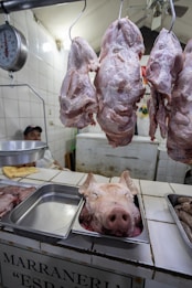 There are several cuts of raw meat hanging on hooks in a butcher shop, with a pig's head displayed in a metal tray on the counter. A person sits in the background partially visible behind a scale.