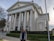 A large neoclassical building with tall white columns and a triangular pediment, featuring banners that read 'YOGA', 'MEDITATION', 'CHANTING', and 'RELAXATION'. The structure is surrounded by neatly trimmed bushes and palm trees. A person stands in the foreground, smiling and wearing casual clothing.