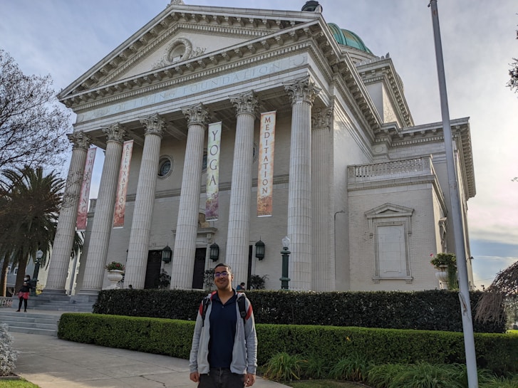 A large neoclassical building with tall white columns and a triangular pediment, featuring banners that read 'YOGA', 'MEDITATION', 'CHANTING', and 'RELAXATION'. The structure is surrounded by neatly trimmed bushes and palm trees. A person stands in the foreground, smiling and wearing casual clothing.