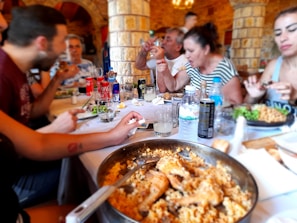 A group of people are gathered around a dining table in what appears to be a rustic-looking restaurant, enjoying a meal together. The table is filled with various types of food and drinks, including a large pan of pasta with seafood, salad, soda cans, and glasses. The people are engaged in conversation and appear to be in a lively, social setting.