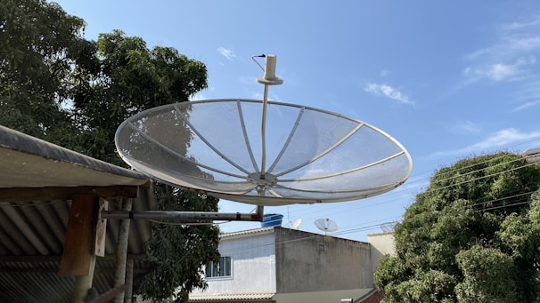 A large satellite dish mounted on a metal bracket is prominently displayed against a clear blue sky. Surrounding it are trees with dense green foliage and a few rooftops of buildings in the background. Additional smaller satellite dishes can be seen on some of the rooftops.