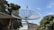 A large satellite dish mounted on a metal bracket is prominently displayed against a clear blue sky. Surrounding it are trees with dense green foliage and a few rooftops of buildings in the background. Additional smaller satellite dishes can be seen on some of the rooftops.