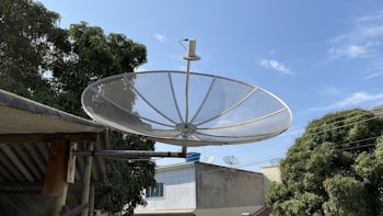 A large satellite dish mounted on a metal bracket is prominently displayed against a clear blue sky. Surrounding it are trees with dense green foliage and a few rooftops of buildings in the background. Additional smaller satellite dishes can be seen on some of the rooftops.