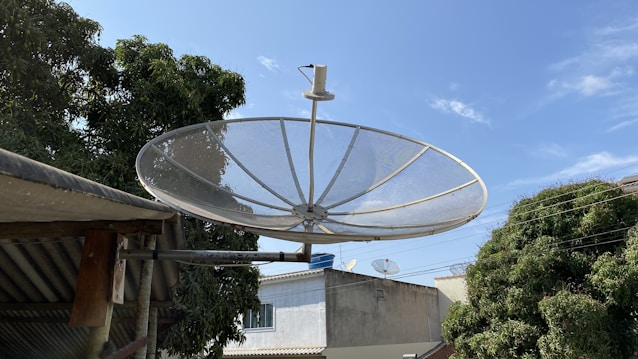 A large satellite dish mounted on a metal bracket is prominently displayed against a clear blue sky. Surrounding it are trees with dense green foliage and a few rooftops of buildings in the background. Additional smaller satellite dishes can be seen on some of the rooftops.