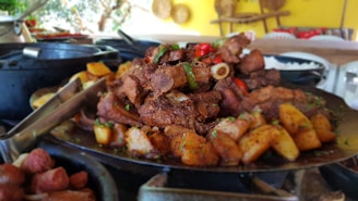 A large platter filled with cooked meat and roasted potatoes is placed on a metal surface. The meat appears to be well-seasoned and garnished with herbs and sliced green peppers. In the background, there are cooking utensils and pots, with bright yellow and warm colors enhancing the ambiance.