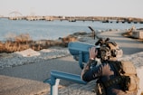 A person wearing tactical gear and a helmet with night vision goggles is looking through a fixed binocular telescope near a waterfront. The scene includes a body of water with a bridge in the background and a pathway lined with rocks.