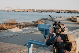 A person wearing tactical gear and a helmet with night vision goggles is looking through a fixed binocular telescope near a waterfront. The scene includes a body of water with a bridge in the background and a pathway lined with rocks.