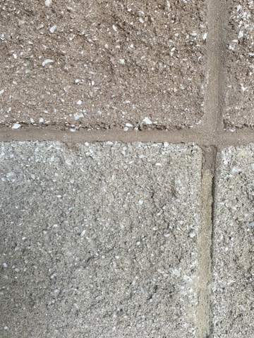A close-up view of a textured concrete block wall. The wall consists of rectangular blocks with a rough surface, featuring small white specks possibly indicating a grainy aggregate. The mortar lines are visible between the blocks, adding structure to the overall appearance.