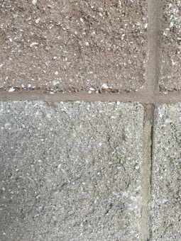 A close-up view of a textured concrete block wall. The wall consists of rectangular blocks with a rough surface, featuring small white specks possibly indicating a grainy aggregate. The mortar lines are visible between the blocks, adding structure to the overall appearance.