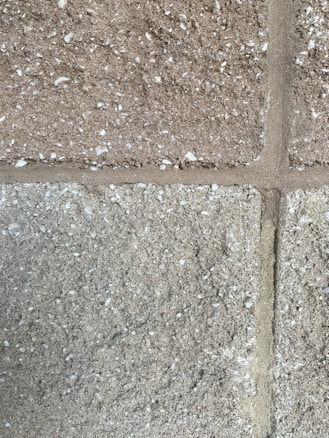 A close-up view of a textured concrete block wall. The wall consists of rectangular blocks with a rough surface, featuring small white specks possibly indicating a grainy aggregate. The mortar lines are visible between the blocks, adding structure to the overall appearance.