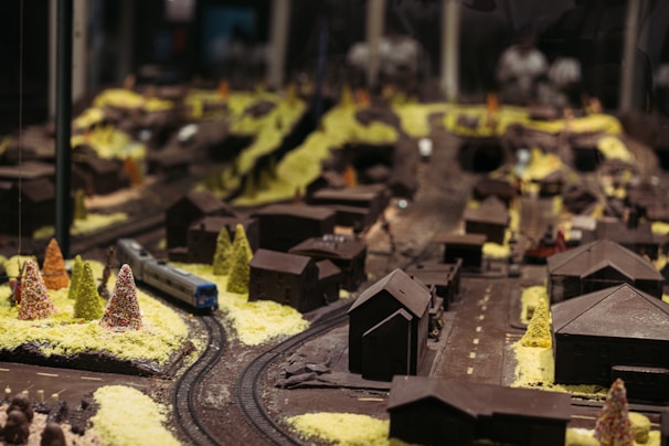 A miniature model of a town featuring numerous small houses, trees, roads, and a train running on tracks. The buildings and landscape are meticulously detailed and simulate a quaint settlement. The scene is softly lit, emphasizing the intricacy of the model's design.