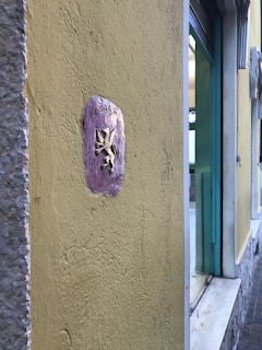 A yellow stucco wall with a purple plaque featuring a winged creature and the words 'CASA' and 'PIA' is visible. The image also shows an open window with a turquoise frame and a view of the interior. The ground is paved with bricks.