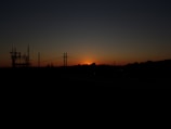 A silhouette of a power station and pylons against a backdrop of a setting sun, with the sky transitioning from orange near the horizon to dark blue at the top.