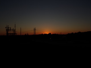 A silhouette of a power station and pylons against a backdrop of a setting sun, with the sky transitioning from orange near the horizon to dark blue at the top.