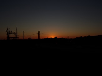 A silhouette of a power station and pylons against a backdrop of a setting sun, with the sky transitioning from orange near the horizon to dark blue at the top.