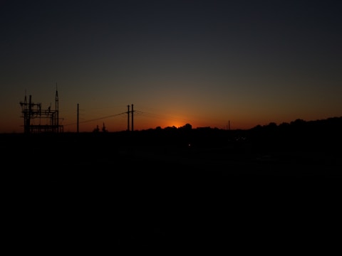 A silhouette of a power station and pylons against a backdrop of a setting sun, with the sky transitioning from orange near the horizon to dark blue at the top.