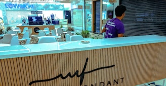 A man wearing a purple shirt is standing behind a counter in a mall. The counter has a wood-paneled design with a logo. In the background, there are several white chairs and tables, and a store with the sign 'LC Waikiki' can be seen. The area appears quiet and unoccupied.