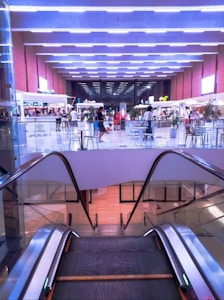 An indoor shopping mall environment featuring an escalator in the foreground leading down to a lower level. The background includes a food court area with several food stands and kiosks, along with tables and chairs where people are seated. Bright, evenly spaced overhead lighting creates a modern and lively atmosphere, while potted plants add a touch of greenery.