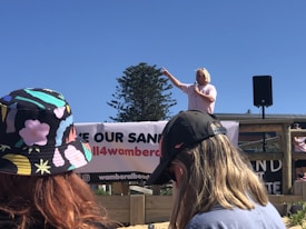 A person speaking into a microphone points to the distance, standing on a platform with a banner that reads 'SAVE OUR SAND.' Two individuals with hats are visible in the foreground, showing their backs to the camera. A loudspeaker stands on the platform, and a tree is seen against the blue sky.