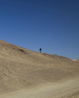 A lone figure walks along the crest of a barren, sandy hill under a clear blue sky, creating a sense of isolation and adventure.