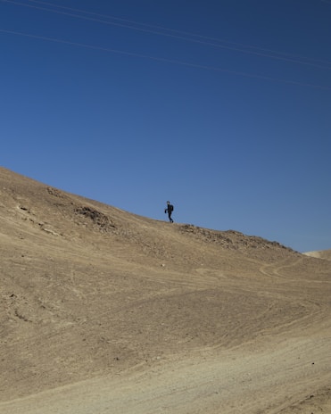 A lone figure walks along the crest of a barren, sandy hill under a clear blue sky, creating a sense of isolation and adventure.