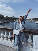 A person wearing a denim jacket and jeans is joyfully posing on a bridge. In the background, the Eiffel Tower is visible across a river, with trees and buildings lining the riverbank. The person has a handbag and appears to be holding a camera.