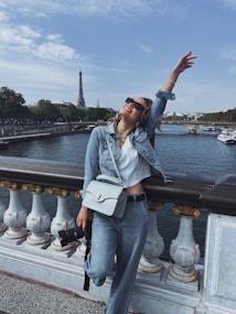 A person wearing a denim jacket and jeans is joyfully posing on a bridge. In the background, the Eiffel Tower is visible across a river, with trees and buildings lining the riverbank. The person has a handbag and appears to be holding a camera.
