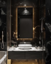 A modern bathroom with a dark marble finish. A rectangular mirror with a backlight hangs above a white oval sink on a marble countertop. Various toiletries are neatly arranged next to the sink. The lighting is subtle, creating a cozy and intimate atmosphere.